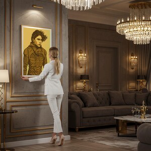 May include: A framed art print featuring a stylized illustration of a person in a brown and yellow outfit. The artwork is displayed in a luxurious room with gold accents, a chandelier, and a woman in a white suit.