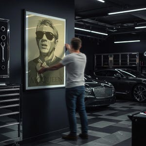 May include: A framed poster featuring a sepia-toned portrait of Steve McQueen wearing sunglasses. The poster has the name "McQUEEN" printed at the bottom. The image is being hung on a dark wall in a garage setting.