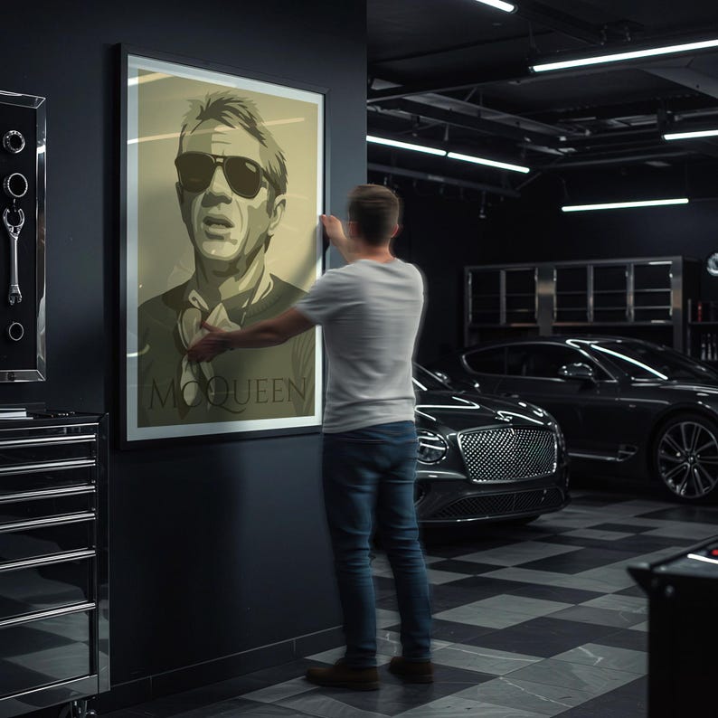 May include: A framed poster featuring a sepia-toned portrait of Steve McQueen wearing sunglasses. The poster has the name "McQUEEN" printed at the bottom. The image is being hung on a dark wall in a garage setting.