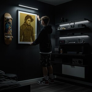 May include: A framed poster featuring a stylized portrait in shades of brown and gold is being hung on a dark wall. A skateboard with a graphic design is mounted on the wall to the left. The room has built-in shelves and LED lighting.