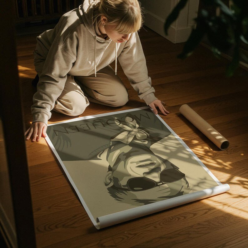 May include: A rolled poster featuring a sepia-toned illustration of a person wearing sunglasses. The poster has the word "MCQUEEN" printed at the top. The poster is on a wooden floor.