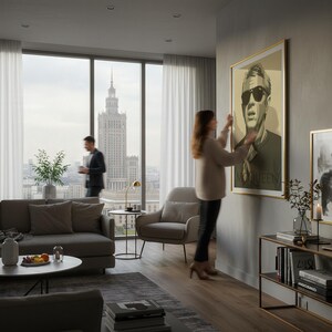 May include: A framed portrait with a gold border hangs on a gray wall. The portrait features a man in sunglasses with the word "McQueen" at the bottom. A woman is adjusting the artwork in a modern living room with a city view.