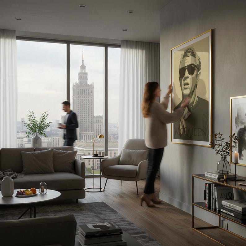 May include: A framed portrait with a gold border hangs on a gray wall. The portrait features a man in sunglasses with the word "McQueen" at the bottom. A woman is adjusting the artwork in a modern living room with a city view.