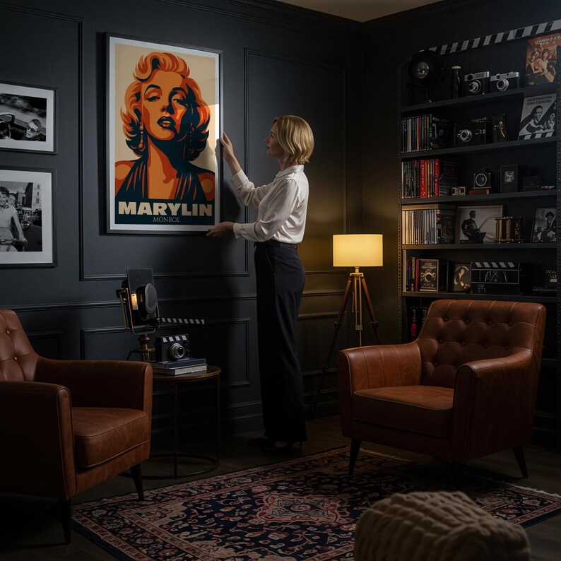 May include: A framed poster featuring a stylized portrait of Marilyn Monroe in shades of orange, blue, and cream. The poster is being hung on a dark wall. The room includes leather armchairs, a patterned rug, and a bookshelf with vintage film equipment.
