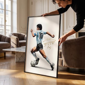 Puede incluir: Ilustración enmarcada de un jugador de fútbol en movimiento, con una camiseta celeste y blanca con el número 10. La obra incluye la firma del jugador y el nombre "DIEGO". El marco es negro.