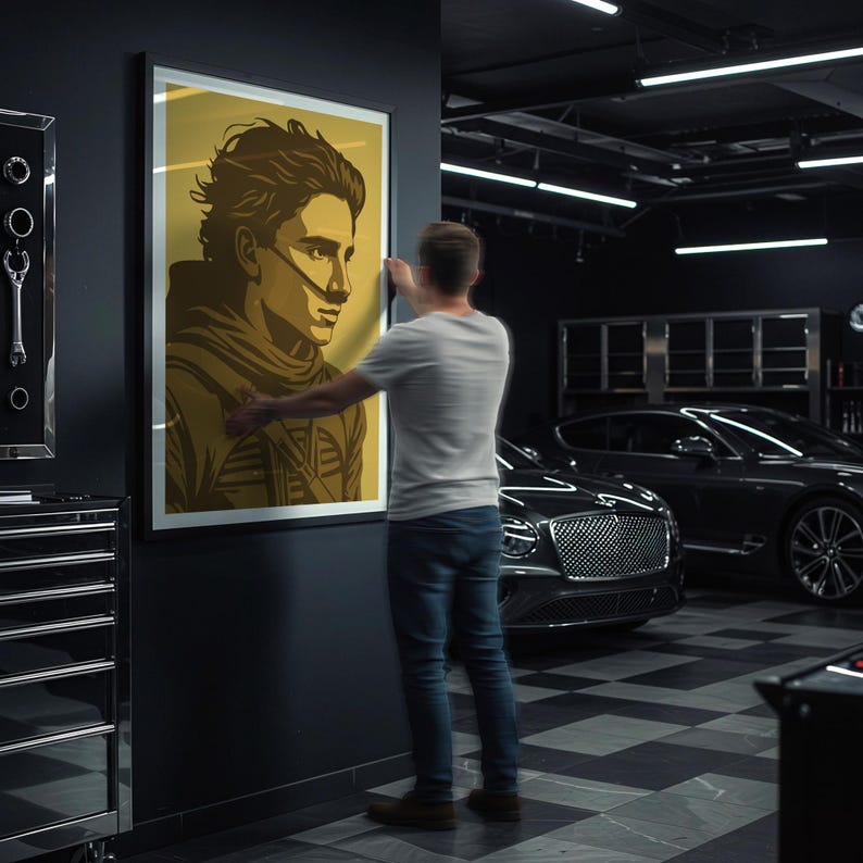 May include: A framed art print featuring a stylized portrait of a person with a neutral expression, in shades of brown and gold. The artwork is being hung on a dark gray wall. The background includes a garage setting with a luxury car.