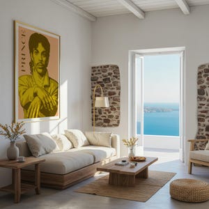 Op de afbeelding: Een woonkamer met een ingelijste poster van Prince, een beige bank en een houten salontafel. De kamer heeft een neutraal kleurenpalet met uitzicht op de oceaan door een open deur. Een gouden lamp en een geweven vloerkleed voegen warmte toe.
