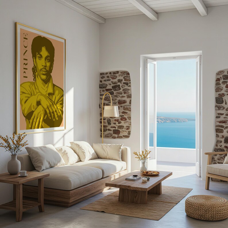 Op de afbeelding: Een woonkamer met een ingelijste poster van Prince, een beige bank en een houten salontafel. De kamer heeft een neutraal kleurenpalet met uitzicht op de oceaan door een open deur. Een gouden lamp en een geweven vloerkleed voegen warmte toe.