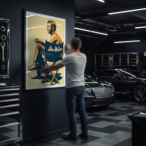 Può includere: Un poster incorniciato con un'illustrazione stilizzata di Steve McQueen su una sedia da regista. Il poster ha uno sfondo color crema con dettagli blu e marroni. Il testo "Steve McQueen" è visibile sulla sedia. Il poster viene appeso a una parete grigio scuro.