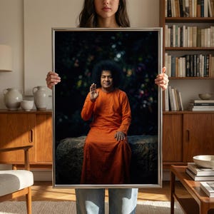 May include: Framed portrait of a man in an orange robe, seated on a stone. The man has dark curly hair and is raising his right hand. The portrait is held by a person in blue jeans.