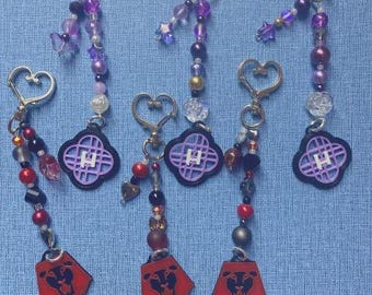 May include: Keychains with heart-shaped clasps and beaded accents in purple, black, and red. Each keychain features a charm with a stylized lion's head and a four-leaf clover design with the letter "H".