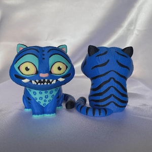 May include: Two blue tiger figurines with black stripes. The front-facing tiger has large yellow eyes, a blue bandana, and a toothy grin. The other tiger is viewed from the back, showing black stripes. The figurines are on a white surface.