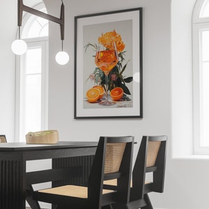 May include: A black framed print of a glass of orange juice with orange slices and flowers. The print is hanging on a white wall above a black dining table with two black chairs.