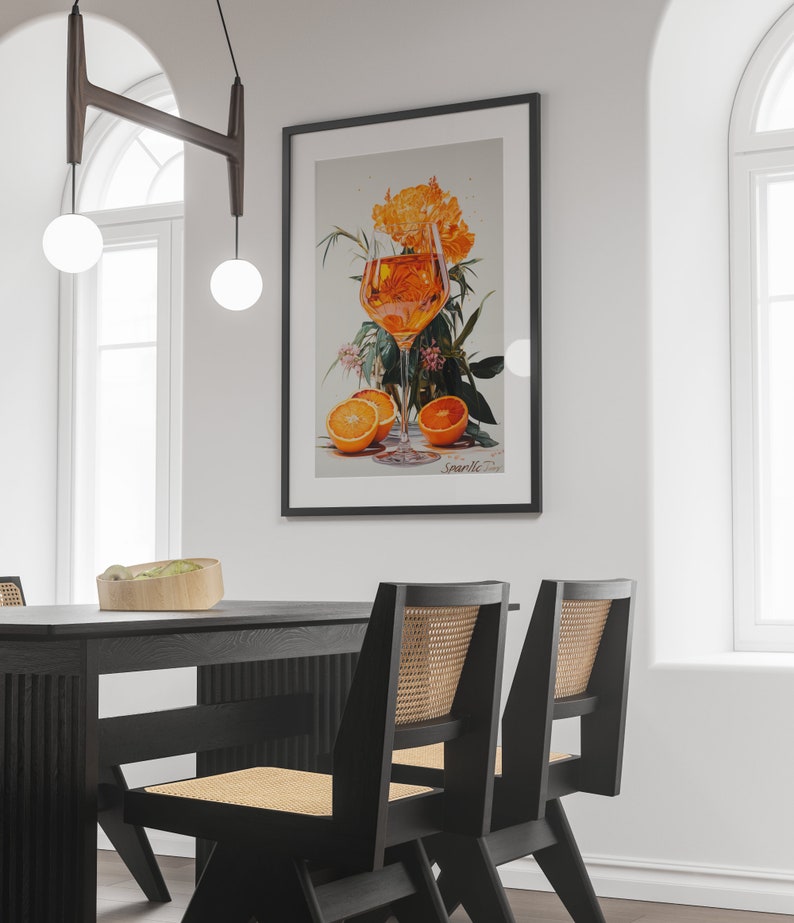 May include: A black framed print of a glass of orange juice with orange slices and flowers. The print is hanging on a white wall above a black dining table with two black chairs.