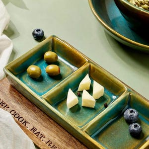 Könnte beinhalten: Ein rechteckiges Serviertablett mit Unterteilungen, glasiert in Türkis und Gold, gefüllt mit Oliven, Käse und Blaubeeren. Das Tablett liegt auf einem Holzschneidebrett mit dem Text "REP. COOK. EAT. REPEAT."