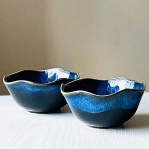 Może przedstawiać: Dwie ceramiczne miski z falistym brzegiem i niebieskim szkliwem gradientowym. Miski przechodzą od ciemnoniebieskiego u podstawy do jasnoniebieskiego u góry, z białym akcentem na krawędzi. Miski są umieszczone na białej powierzchni.
