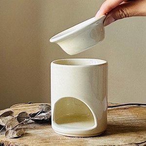 Op de afbeelding: Een crèmekleurige keramische oliebrander met een gebogen opening en een bijpassend deksel. De brander staat op een houten oppervlak, met een hand die het deksel erboven houdt. Het ontwerp is eenvoudig en modern.