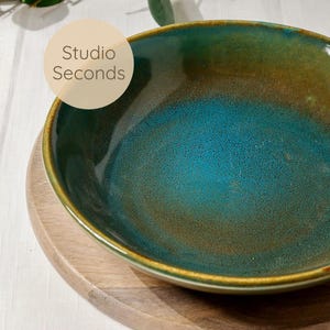 May include: A ceramic bowl with a teal and brown glaze, set on a wooden plate. The bowl has a glossy finish and a wide rim. The text "Studio Seconds" is in a white circle in the upper left corner. The bowl is likely for serving food.