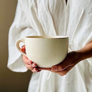 May include: Cream-colored ceramic mug with a large handle, speckled texture, and a brown rim. The mug is held in both hands, suggesting a comfortable size for beverages. The overall aesthetic is rustic and minimalist.