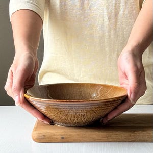 May include: A brown ceramic bowl with a textured surface is held by a person. The bowl has a wide, shallow shape with a slightly raised rim. The bowl is sitting on a wooden cutting board.