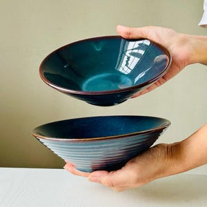 May include: Two ceramic bowls, one stacked on top of the other. The top bowl is a deep teal color with a dark brown rim. The bottom bowl is a dark blue color with a dark brown rim. Both bowls have a subtle ribbing pattern.