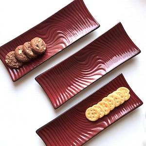 Peut inclure: Trois plateaux de service rectangulaires bordeaux avec une texture ondulée et des bordures sombres. L'un contient des cookies aux pépites de chocolat, un autre des crackers. Les plateaux sont disposés en diagonale.