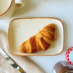 Può includere: Un croissant dorato e croccante è appoggiato su un piatto rettangolare in ceramica macchiata con bordo marrone. Sono visibili un tovagliolo di maglia beige e un coltello d'argento. Una tazza e un barattolo di crema spalmabile al cioccolato sono anch'essi nell'immagine.
