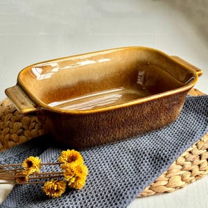 Könnte beinhalten: Eine rechteckige, braune Keramik-Backform mit Griffen. Die Form hat eine glänzende Oberfläche und steht auf einem grauen Tuch und einer gewebten Matte. Gelbe Blumen sind vor der Form platziert.