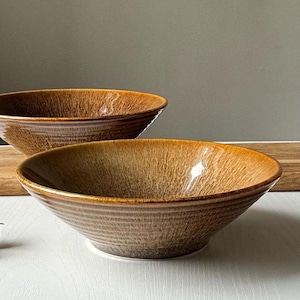 May include: Two brown ceramic bowls with a textured, earthy design. The bowls have a wide, shallow shape and a glossy finish. The bowls are suitable for serving soups, salads, or other dishes. The bowls are placed on a white surface.