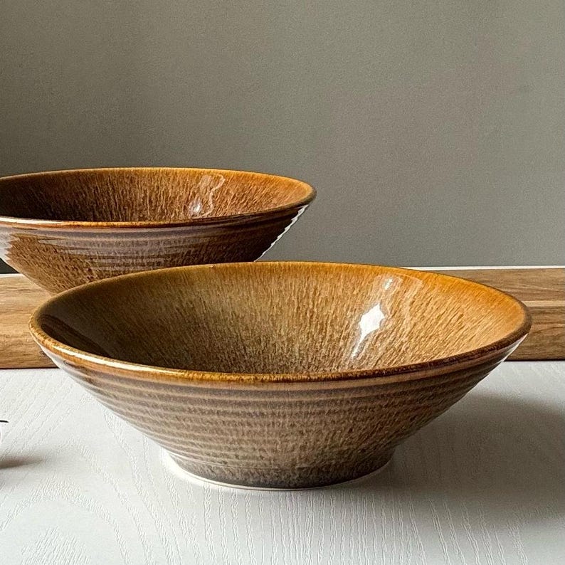 May include: Two brown ceramic bowls with a textured, earthy design. The bowls have a wide, shallow shape and a glossy finish. The bowls are suitable for serving soups, salads, or other dishes. The bowls are placed on a white surface.