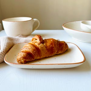 May include: A golden-brown croissant rests on a speckled, rectangular plate with a brown rim. A white mug and a bowl are in the background. The scene suggests a breakfast or brunch setting.