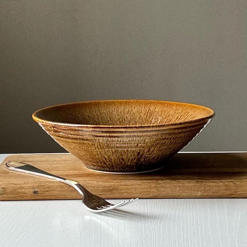 May include: A brown ceramic bowl with a textured surface and a silver fork on a wooden cutting board. The bowl has a wide rim and a slightly flared shape. The fork is placed to the left of the bowl.