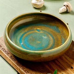 Op de afbeelding: Een keramische kom met een glanzende afwerking, met een mix van turquoise, goud en bruin tinten. De binnenkant van de kom toont een wervelend patroon van turquoise en goud. De rand is een donkerdere bruine tint.