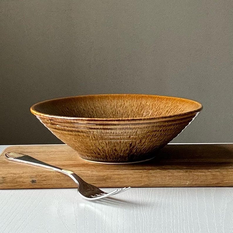 May include: A brown ceramic bowl with a textured surface. The bowl is sitting on a wooden cutting board with a silver fork.
