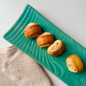 Può includere: Quattro pasticcini dorati con un motivo a spirale sono disposti su un piatto da portata verde acqua a forma di foglia. Un tovagliolo beige strutturato è parzialmente visibile nell'angolo in basso a sinistra. I pasticcini hanno un ripieno chiaro.