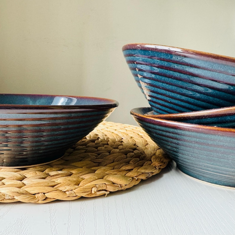 May include: A stack of four blue ceramic bowls with a textured, ridged design. The bowls have a dark brown rim and sit on a woven, beige placemat. The bowls are on a white surface.