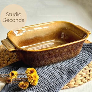 May include: A rectangular, brown ceramic baking dish with handles. The dish has a glossy finish and is set on a blue cloth and woven mat. The text "Studio Seconds" is in a circle in the upper left corner.
