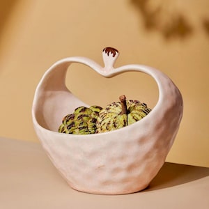 May include: A light pink ceramic fruit bowl with a heart-shaped handle and a brown accent. The bowl contains two green and yellow custard apples. The bowl has a textured surface and is on a light-colored surface.