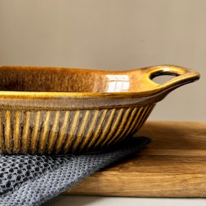 May include: A brown ceramic baking dish with a ribbed exterior and a handle with a hole. The dish is set on a wooden chopping board and a grey cloth. The dish has a glossy finish.