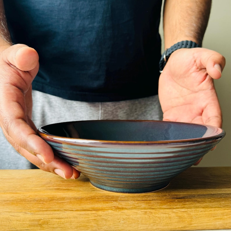 May include: A dark blue ceramic bowl with a textured, striped design. The bowl is empty and sitting on a wooden surface.