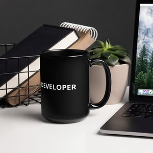 以下が含まれることがあります： 白い「DEVELOPER」の文字が書かれた黒いセラミック製のコーヒーカップ。カップは、ラップトップ、鉢植えの多肉植物、ノートの束の隣にある白い表面の上に置かれています。