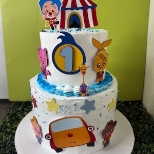 May include: A three-tiered white cake decorated with cartoon characters and a circus tent topper. The cake features blue frosting accents, star-shaped sprinkles, and a large number "1" on the second tier, suggesting a first birthday celebration.