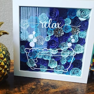Puede incluir: Una caja de sombra con marco blanco con un arreglo floral en tonos azules. El diseño incluye un contorno de bañera y la palabra "relax". Las flores son de papel, creando un efecto 3D.