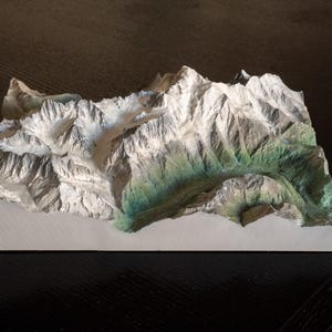 以下が含まれることがあります： 山脈の3Dモデル。白とグレーの山頂と緑と茶色の谷があります。長方形の明るい色のベースに取り付けられており、地形を詳細に示しています。