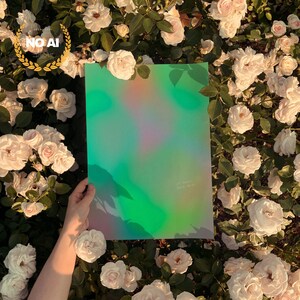 May include: A hand holds a rectangular, iridescent green sheet with a soft, colorful pattern. The sheet is surrounded by white roses and green foliage. The text "LET YOUR DREAMS BLOOM" is visible on the sheet.