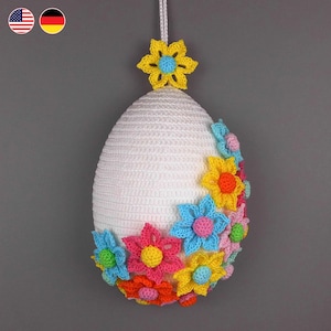 Könnte beinhalten: Weißes gehäkeltes Osterei-Ornament, verziert mit bunten gehäkelten Blumen. Das Ei hängt an einer weißen Schnur.
