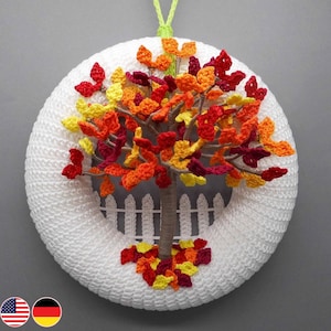 Könnte beinhalten: Ein weißer gehäkelter Kranz mit einem herbstlichen Baum in der Mitte. Der Baum hat braune Äste und Blätter in Rot-, Orange- und Gelbtönen. Der Kranz ist mit einem weißen Lattenzaun und einem kleinen Blätterhaufen am Fuß des Baumes verziert.