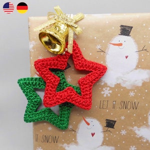 Könnte beinhalten: Zwei gehäkelte Sterne, einer rot und einer grün, sind an braunem Geschenkpapier mit einer goldenen Glocke befestigt. Das Geschenkpapier hat ein Schneemann-Design und den Text "Let it snow".