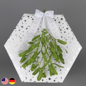 Könnte beinhalten: Eine sechseckige Geschenkbox, verpackt in weißem Papier mit silbernen Sternen, verziert mit grünem gehäkeltem Mistelzweig und einem weißen Band mit der Aufschrift "MERRY CHRISTMAS". Der Mistelzweig ist mit kleinen weißen Perlen verziert.