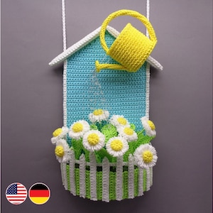 Può includere: Appeso a parete lavorato a uncinetto con una casa blu con un annaffiatoio giallo che versa acqua su una staccionata bianca con margherite bianche.